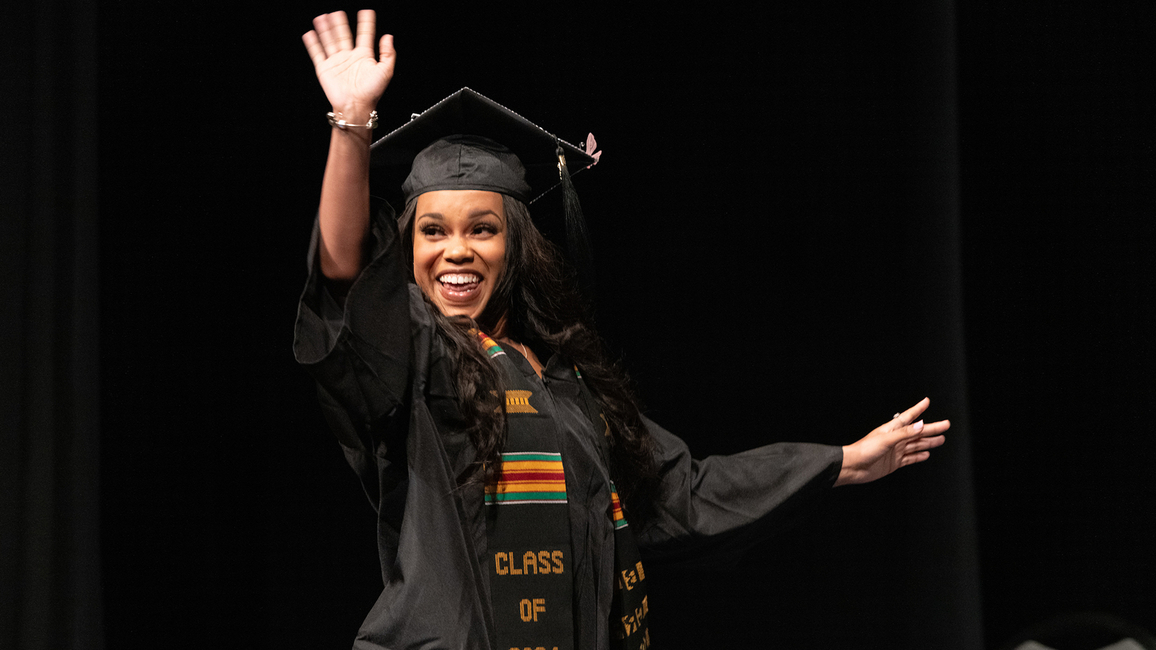 A female UIS graduate waves to the crowd in her cap and gown while walking on stage to collect her degree.