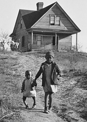 A black and white photo showing a house on top of a hill with two children, one with Rickets, walking towards the camera. 