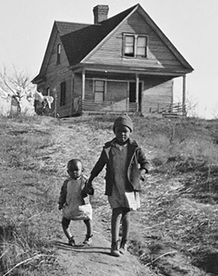 A black and white photo showing a house on top of a hill with two children, one with Rickets, walking towards the camera. 