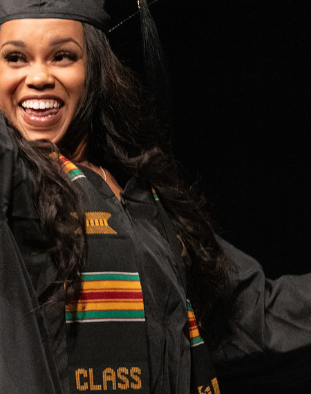 A female UIS graduate waves to the crowd in her cap and gown while walking on stage to collect her degree.