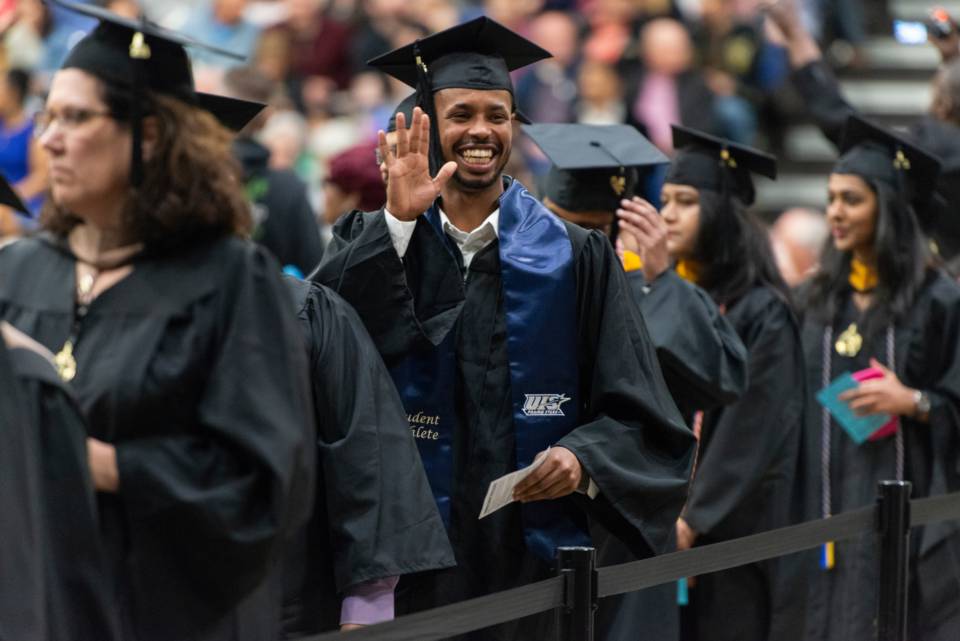 Graduate waves and smiles while standing in a line of other graduates