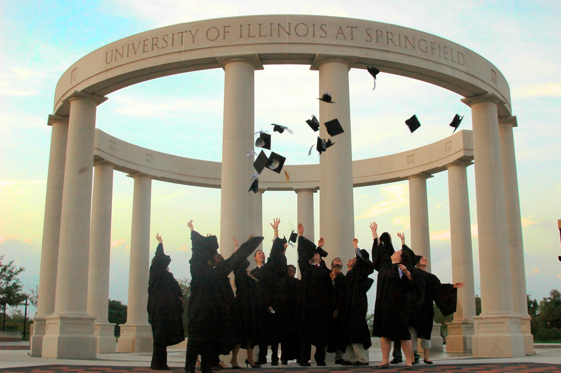 A group of graduates toss their graduation caps into the air in front of the UIS colonnade