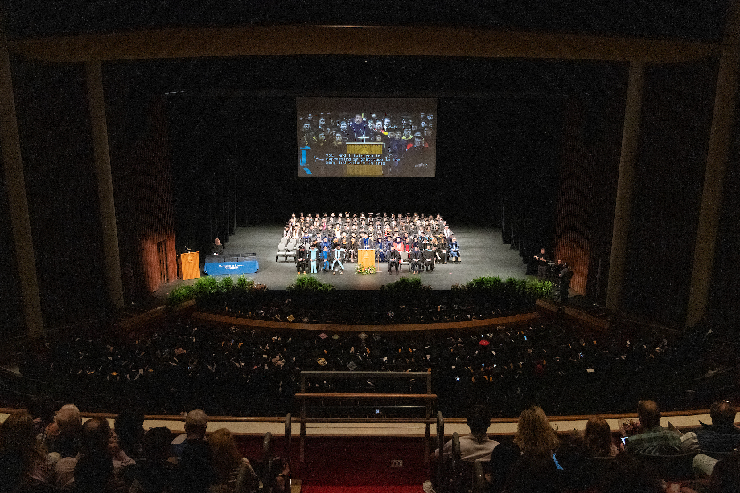 A view of the College of Health, Science and Technology graduates seated on stage in regalia on the stage of the Sangamon Auditorium with the audience in the foreground.