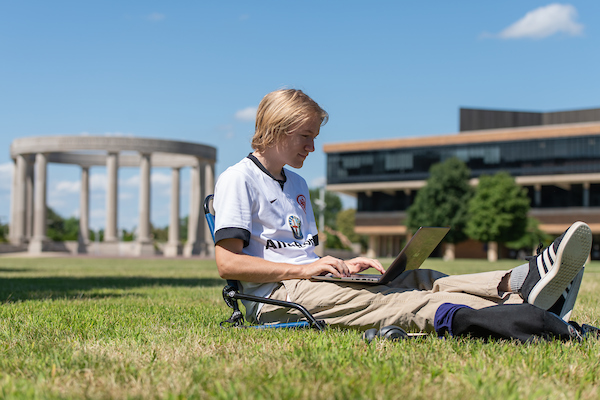 UIS student studying on a laptop outdoors with the colonnade and library in the background on a sunny day