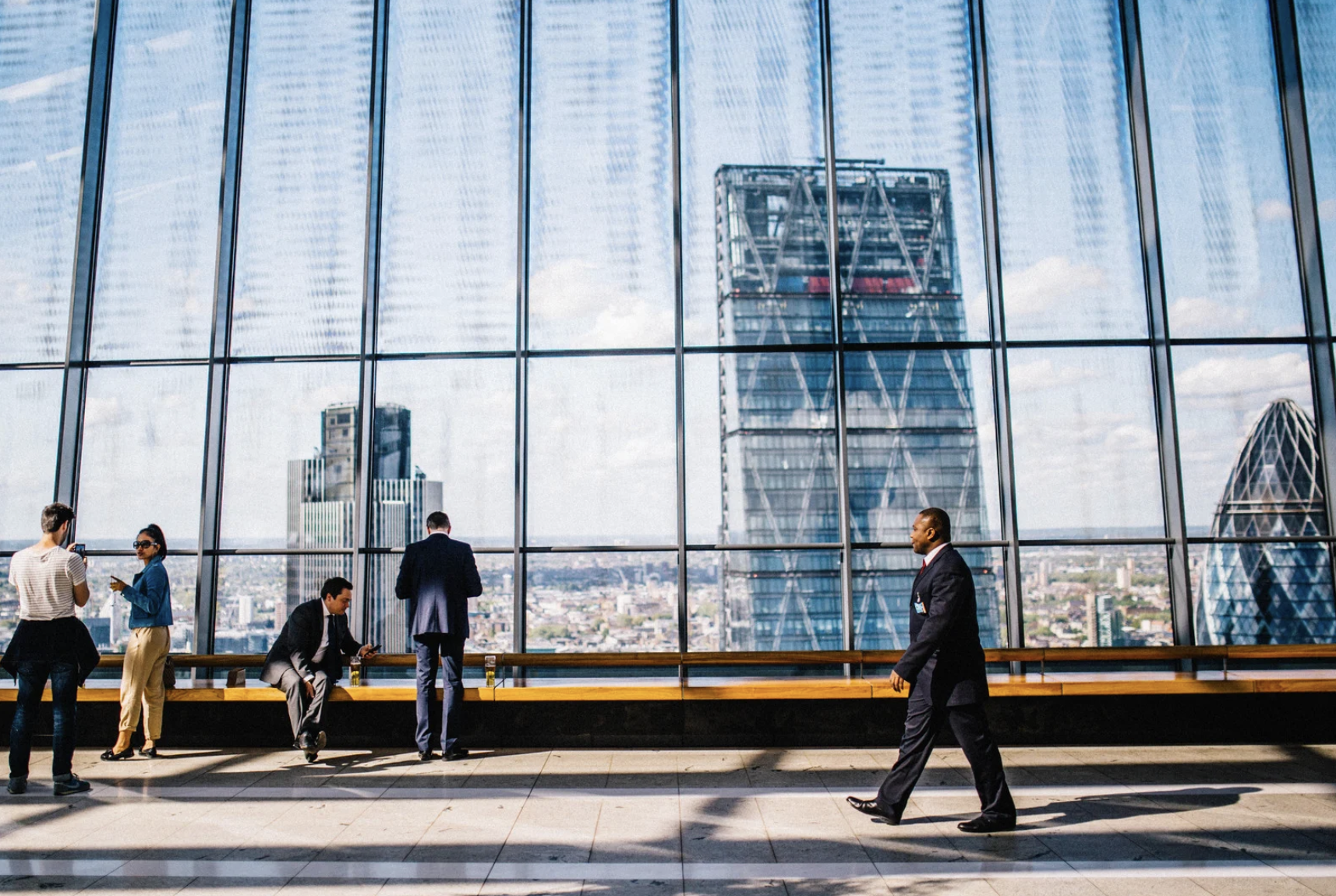 Man walking on sidewalk near people standing and sitting beside curtain-wall building 