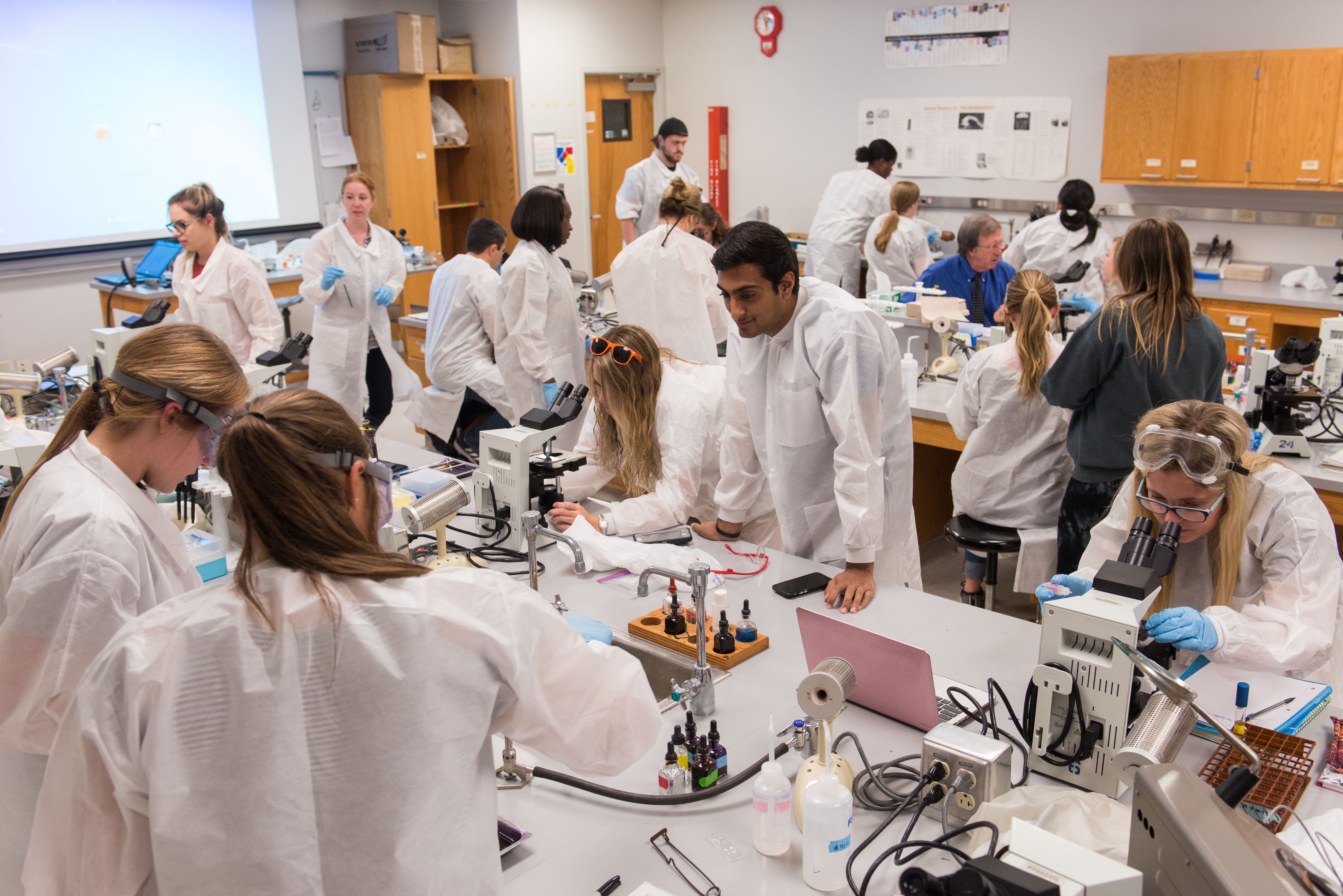 Students in lab coats work with microscopes and scientific instruments in the biology laboratory during a hands-on class session