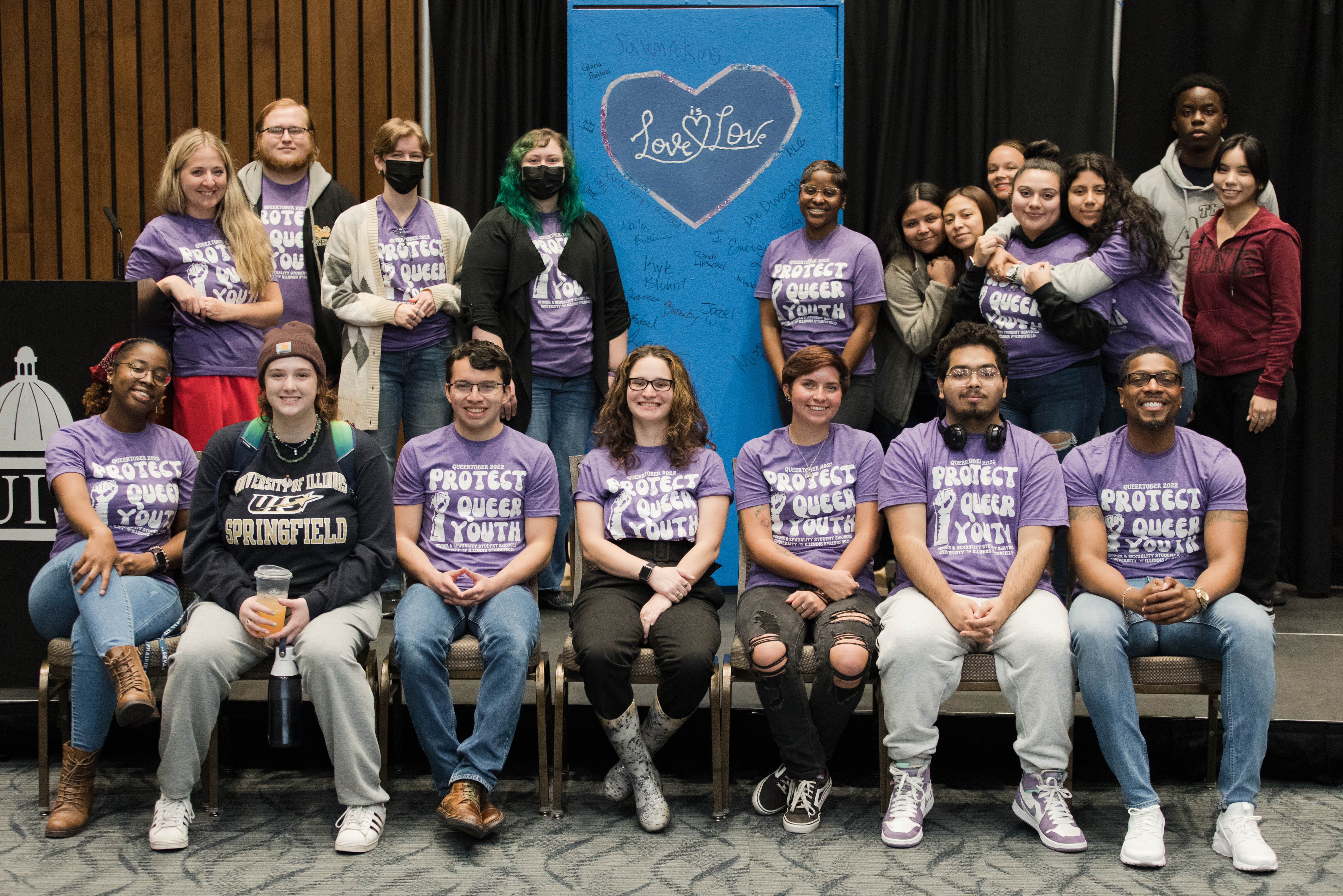 People sitting and standing on a stage, posing with a door that says "Love is Love".