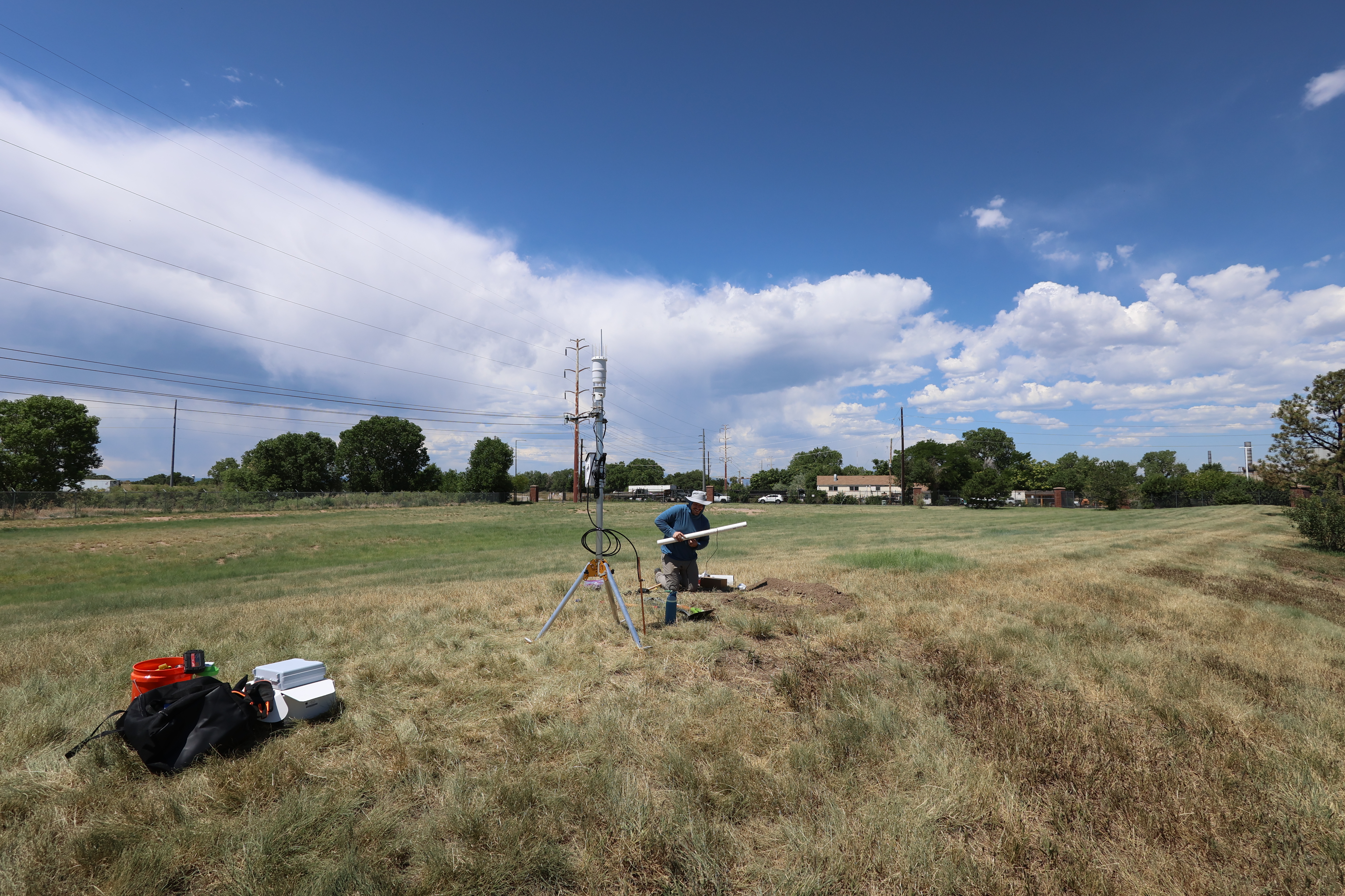 Researcher installing soil moisture and weather monitoring equipment in an open grass field under a clear sky