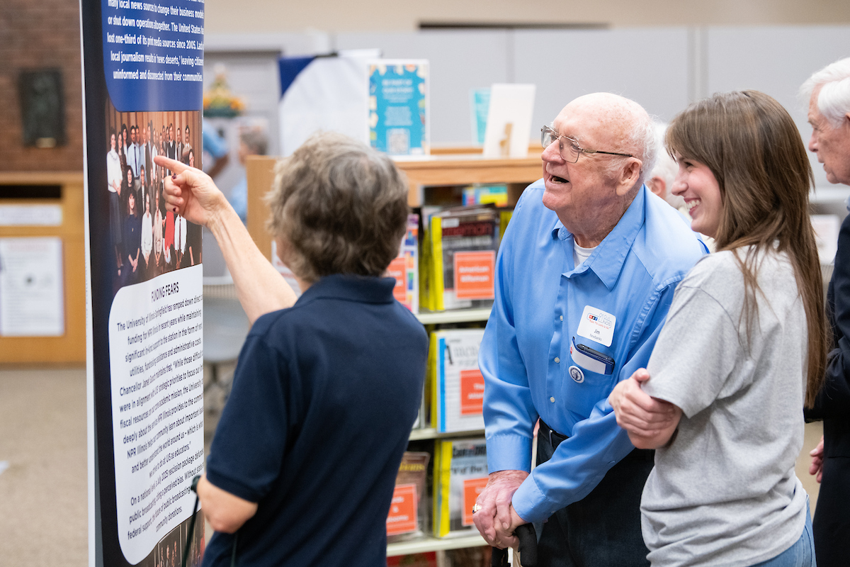 Older man and younger woman smiling, reading a display in a library setting., Randy Eccles addresses a large crowd from a staircase landing inside a building., Ribbon-cutting opening the new exhibit Radio that Listens to you 50 years of NPR Illinois, Jim Newbanks and Evie Rodenbaugh take a look at the exhibit Radio that Listens to You