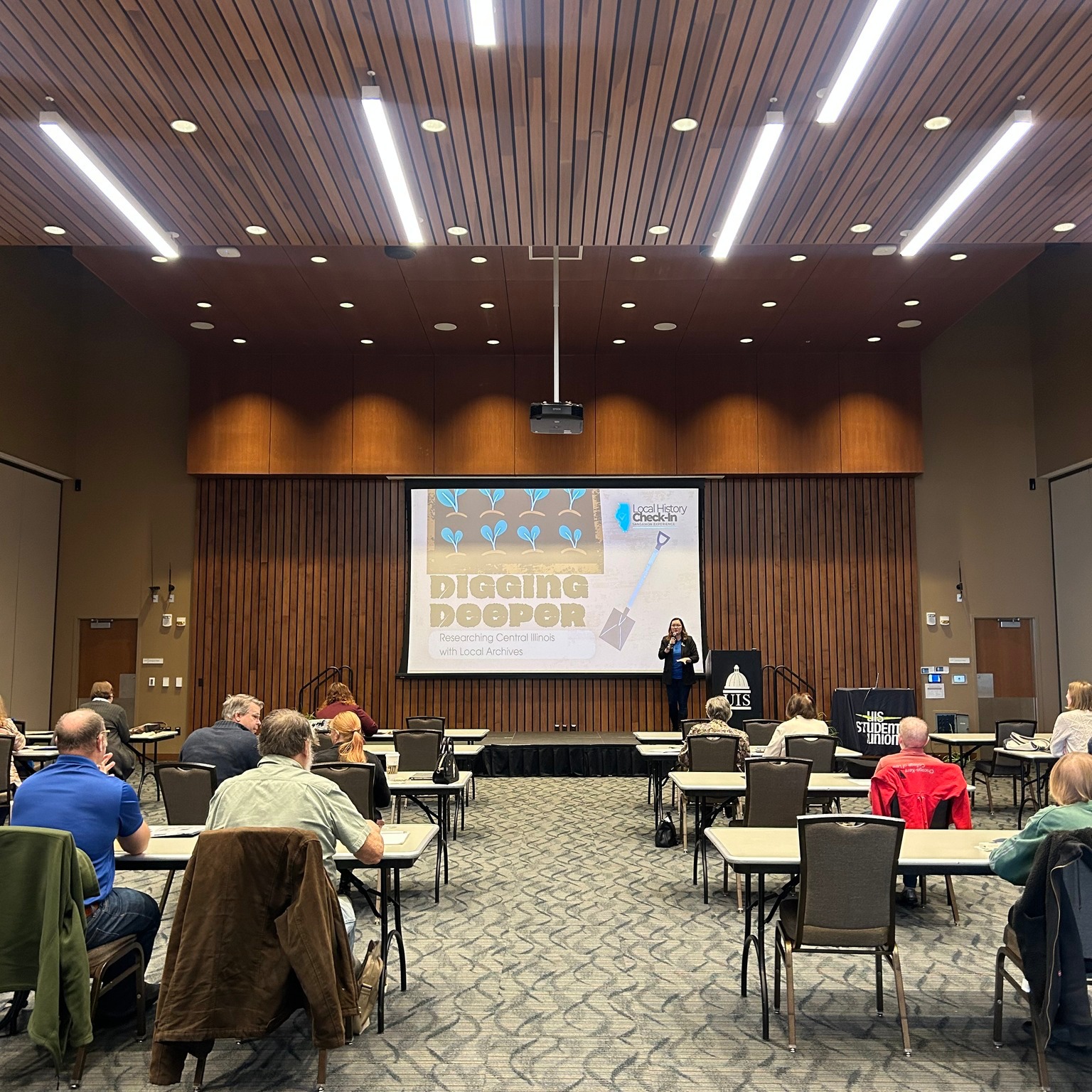 A conference room with Local History Check-In attendees seated at tables facing a presentation screen.