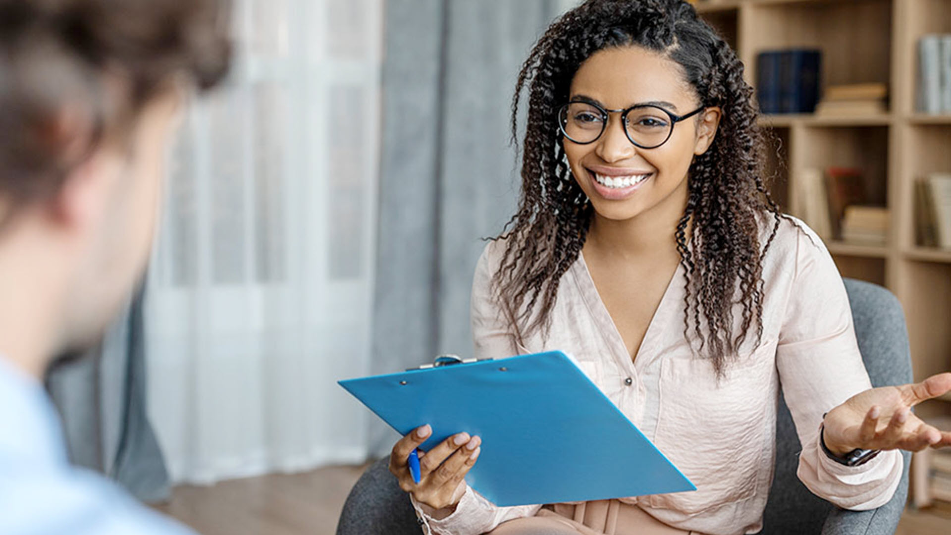 Woman with glasses smiling, holding clipboard, sitting across from another person.
