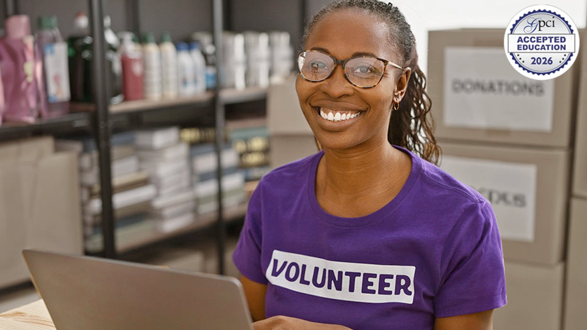 Smiling volunteer in a purple shirt, working on a laptop in a storage room.