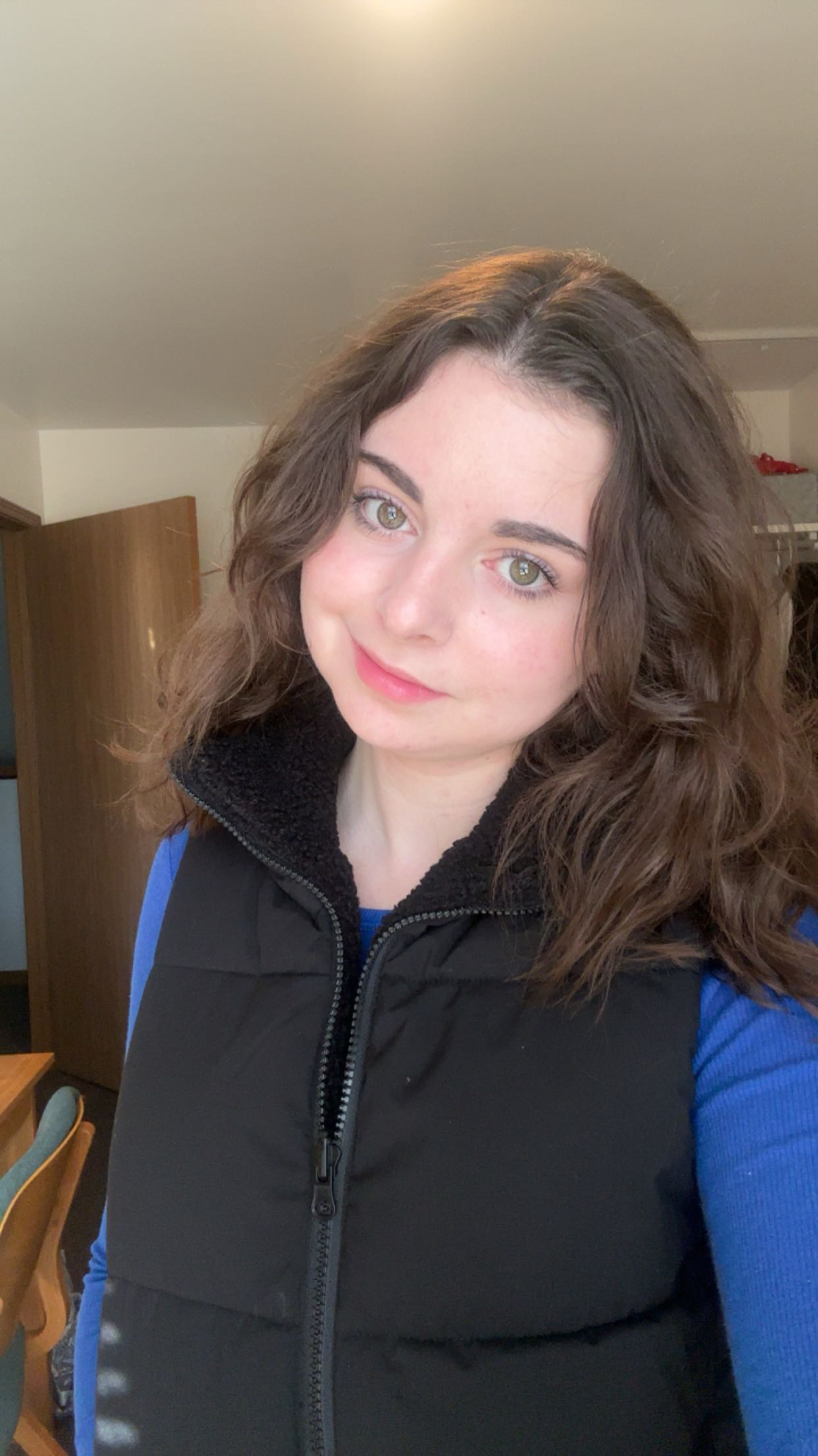 Smiling person with wavy hair in a blue top and black vest indoors.