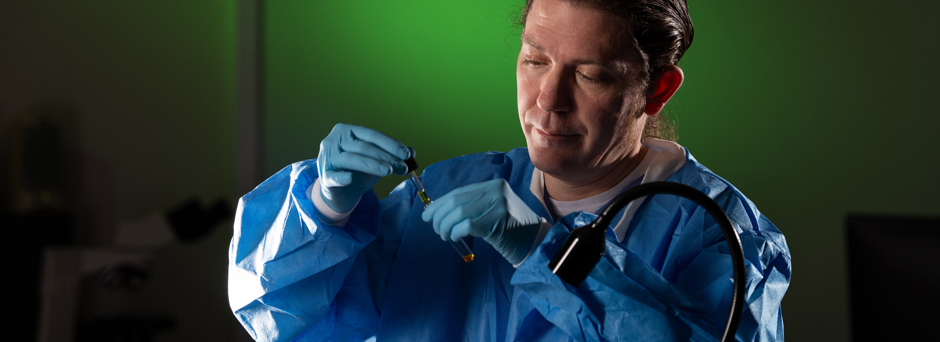 Scientist in blue protective gear under green lighting using a dropped to add liquid in beaker.