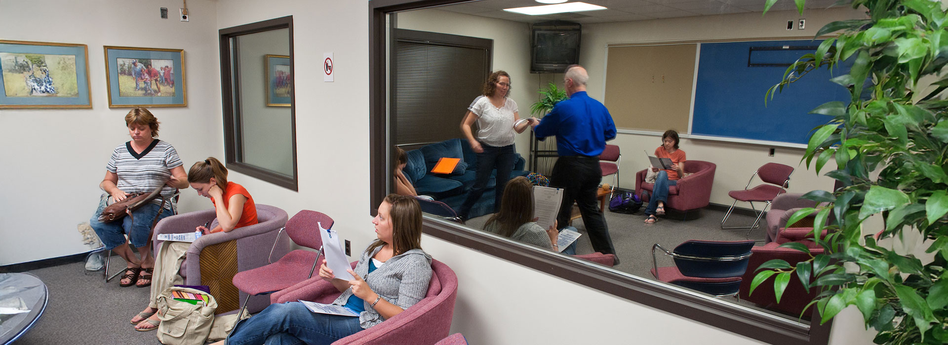 People sitting in a waiting room with red chairs and paintings on the wall.