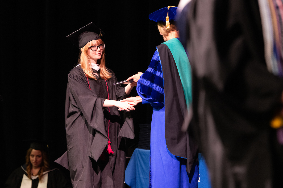 Graduate receives diploma on stage from faculty member in blue gown.