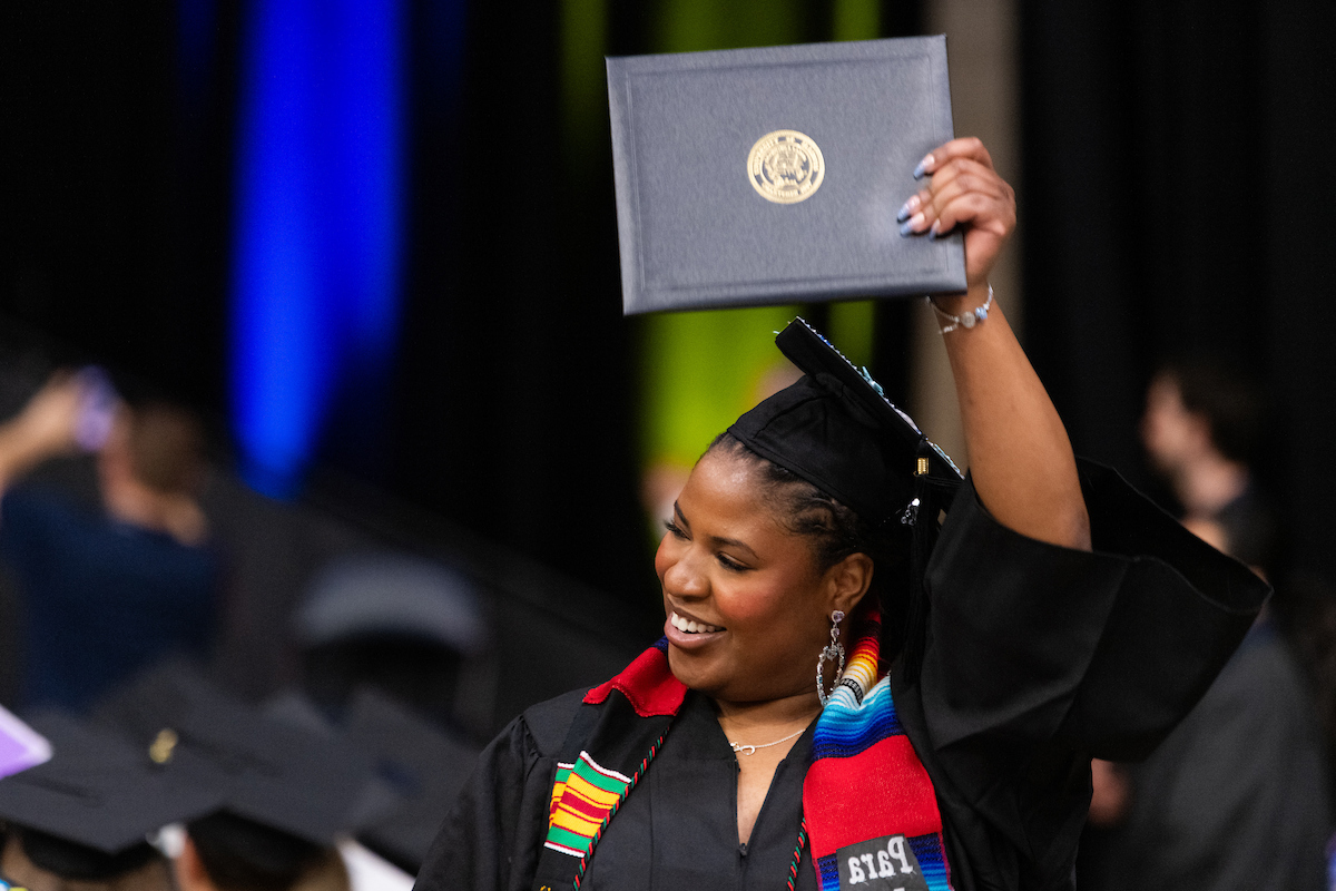 Graduate joyfully holding diploma at ceremony, wearing colorful graduation stole.
