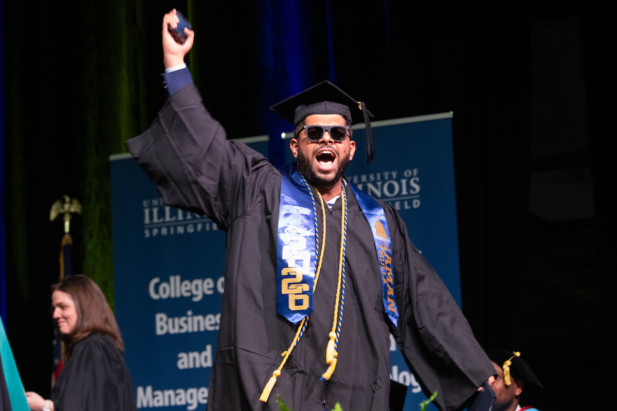 Graduate celebrating on stage in dark hall, wearing cap and blue stole.