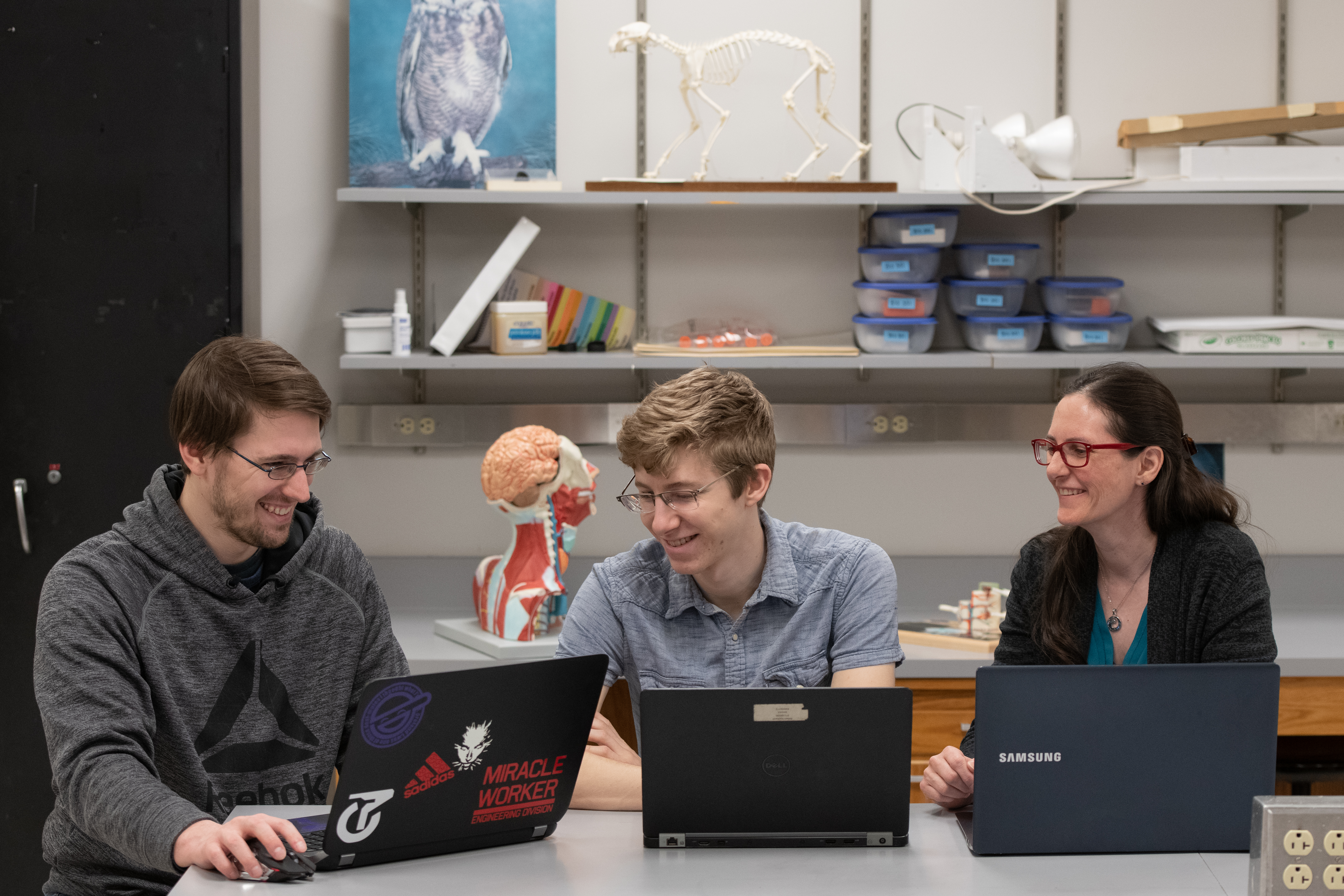 Three people smiling, working on laptops in a lab setting.