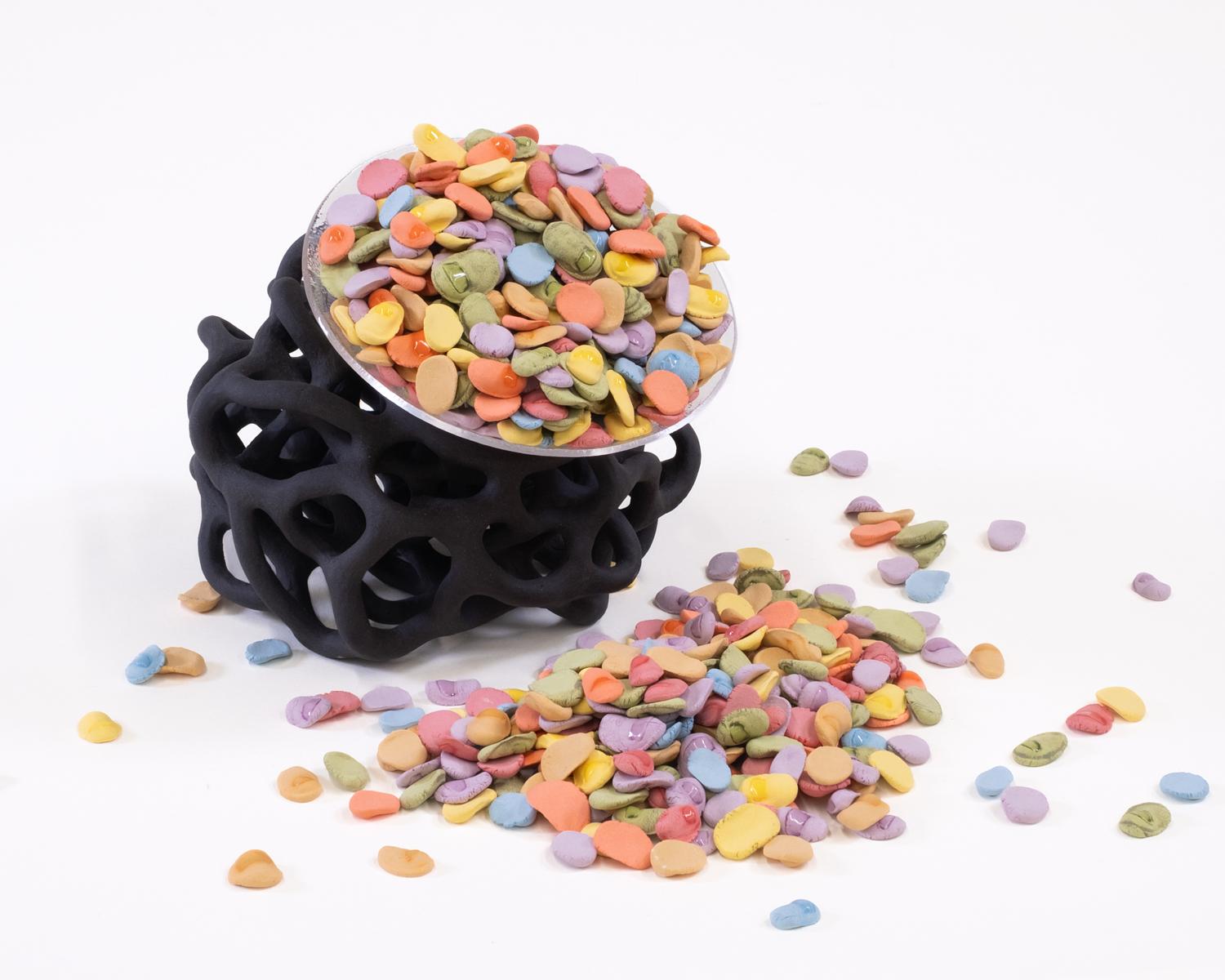A ceramic and glass vessel that holds a mound of pastel, pebble-like pieces with many of the pieces have spilled onto the white surface around the artwork