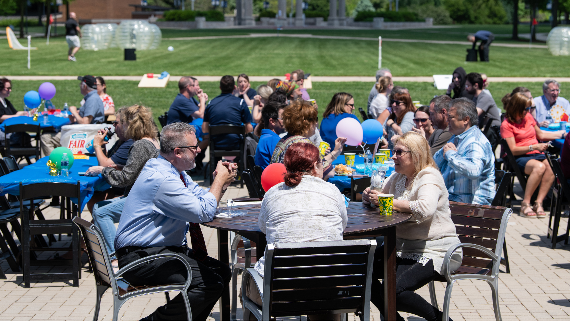 Outdoor gathering with people seated at tables, balloons, and sunny weather.