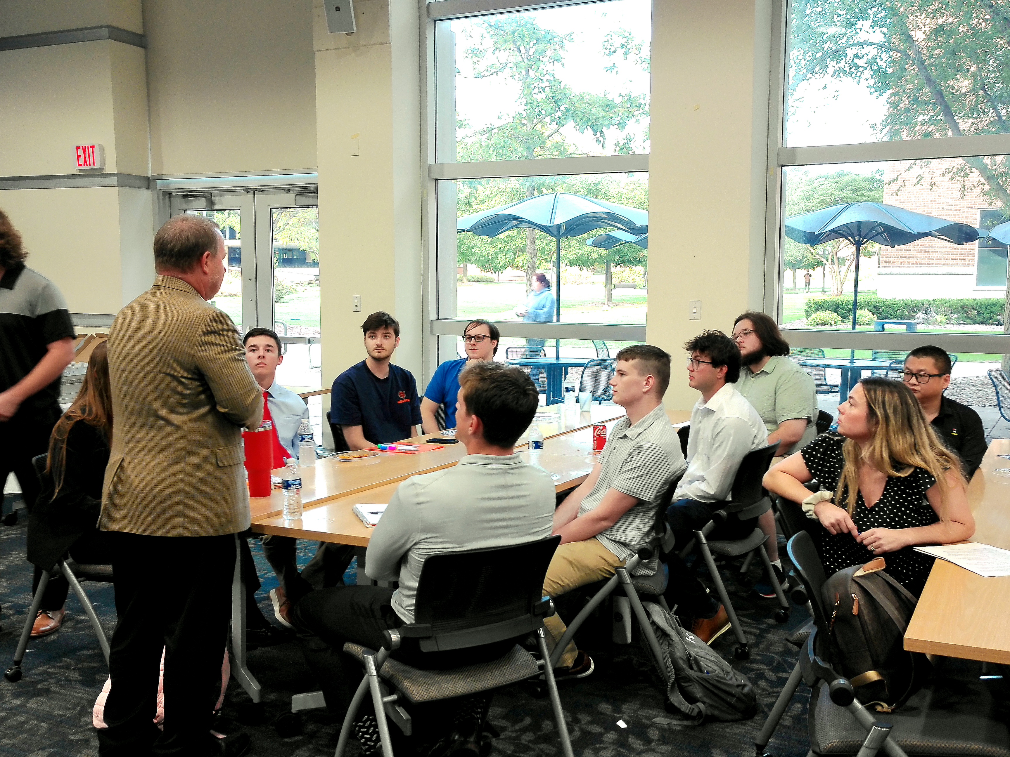 A group of people sit around a table listening to a speaker in a bright room.