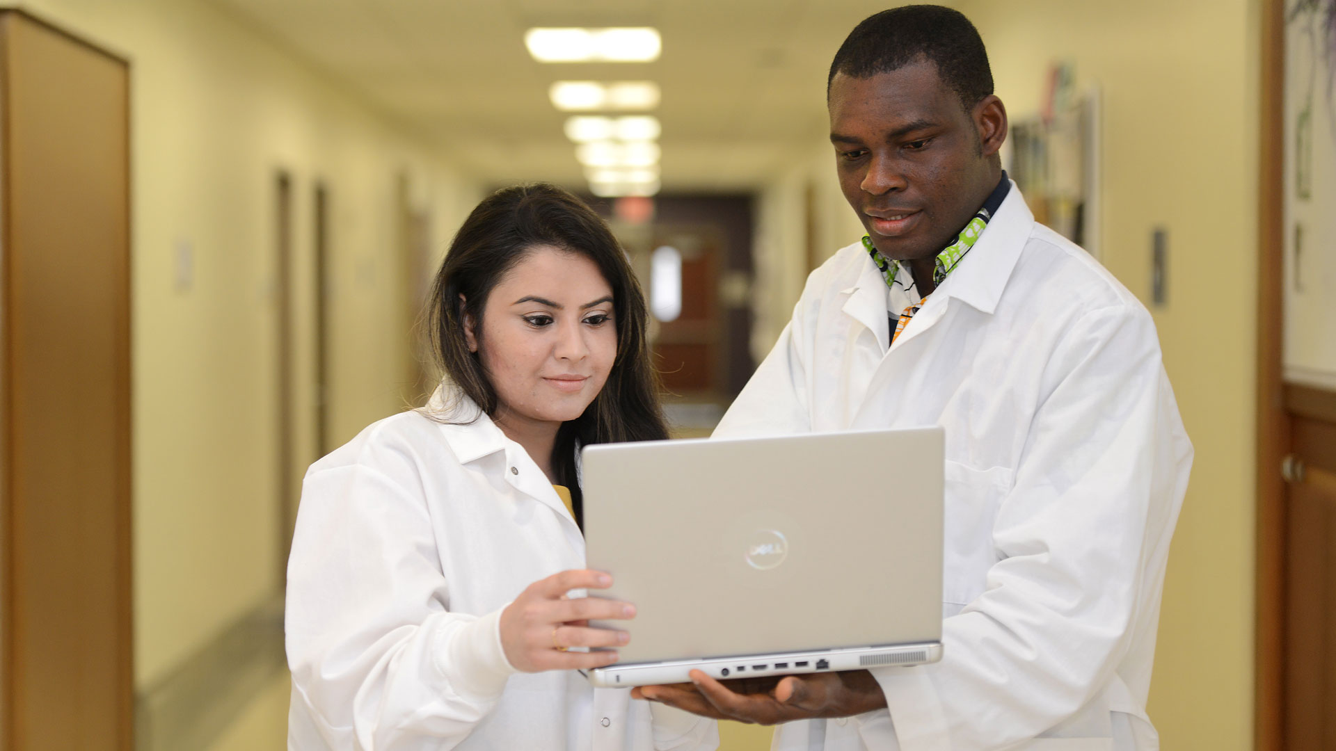 two healthcare informatics students in lab coats looking at a laptop in a hallway