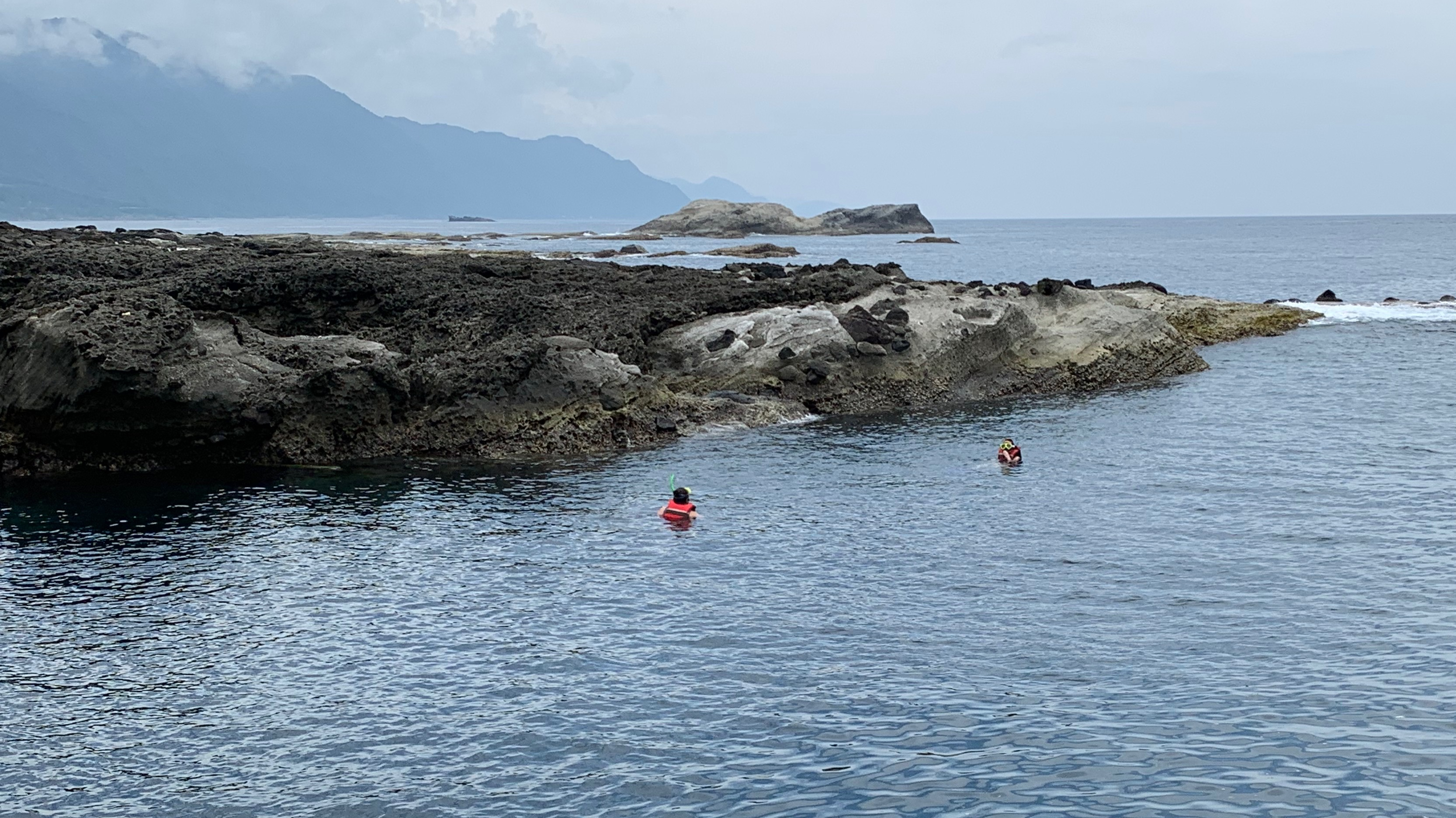 Two students are in the ocean along a rocky and mountainous coast.
