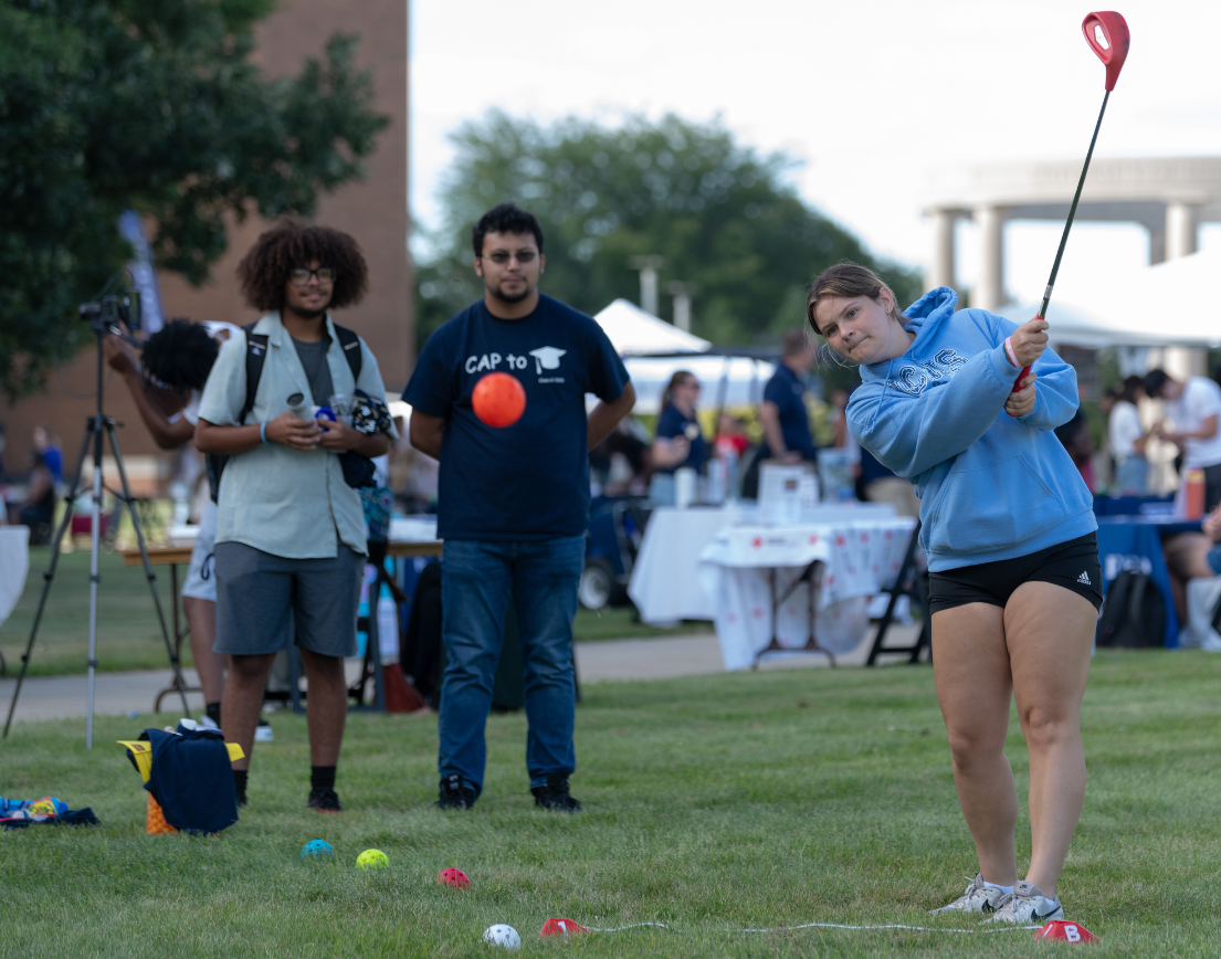 Three students stand outside on the grass. One student swings a club after hitting a ball while the other two watch.