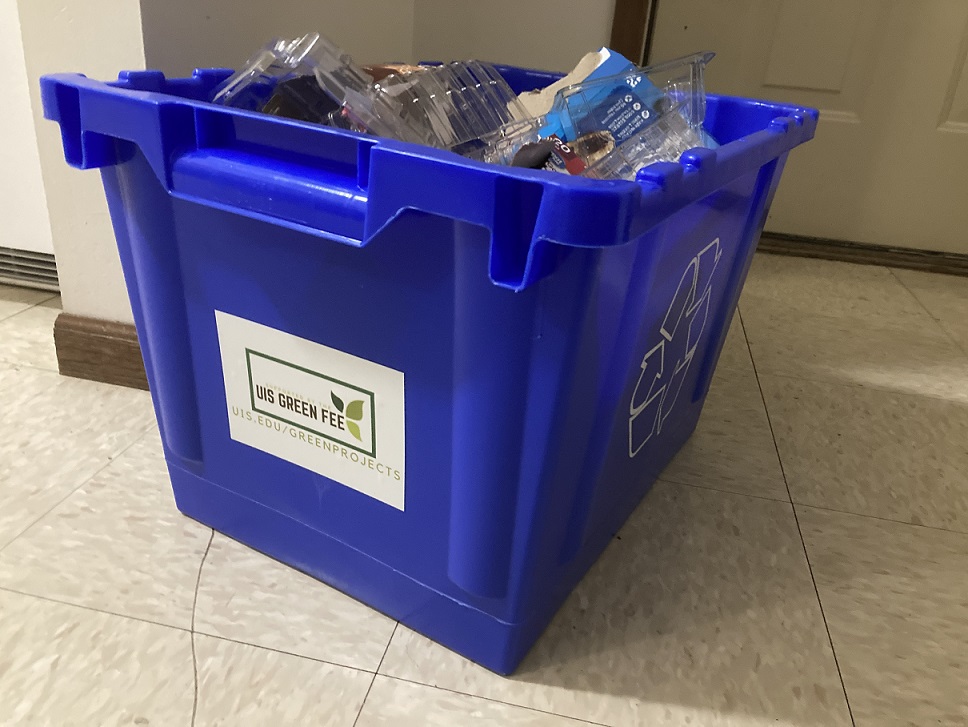 A filled recycling bin, with a sticker that has the Green Fee Commitee logo facing the camera.