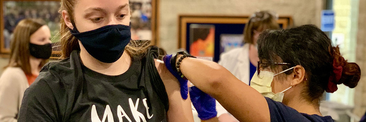 Student in a mask receiving a flu shot from another masked individual.