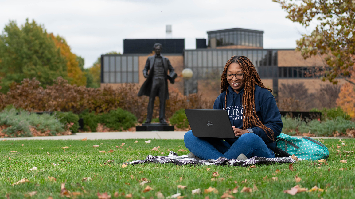 A student sitting on the grass on the UIS quad with a laptop computer with a statue of Abraham Lincoln and the HSB Building in the background.