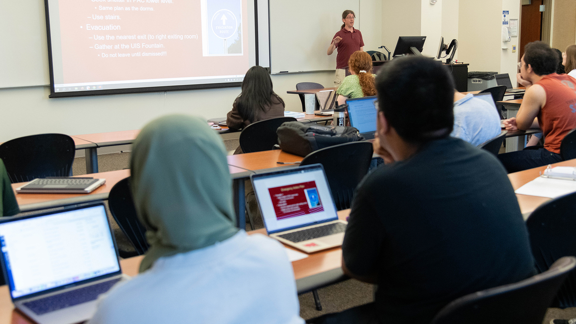 The backs of heads of students sitting in a classroom listing to a professor teach. Several laptop computers are visible.