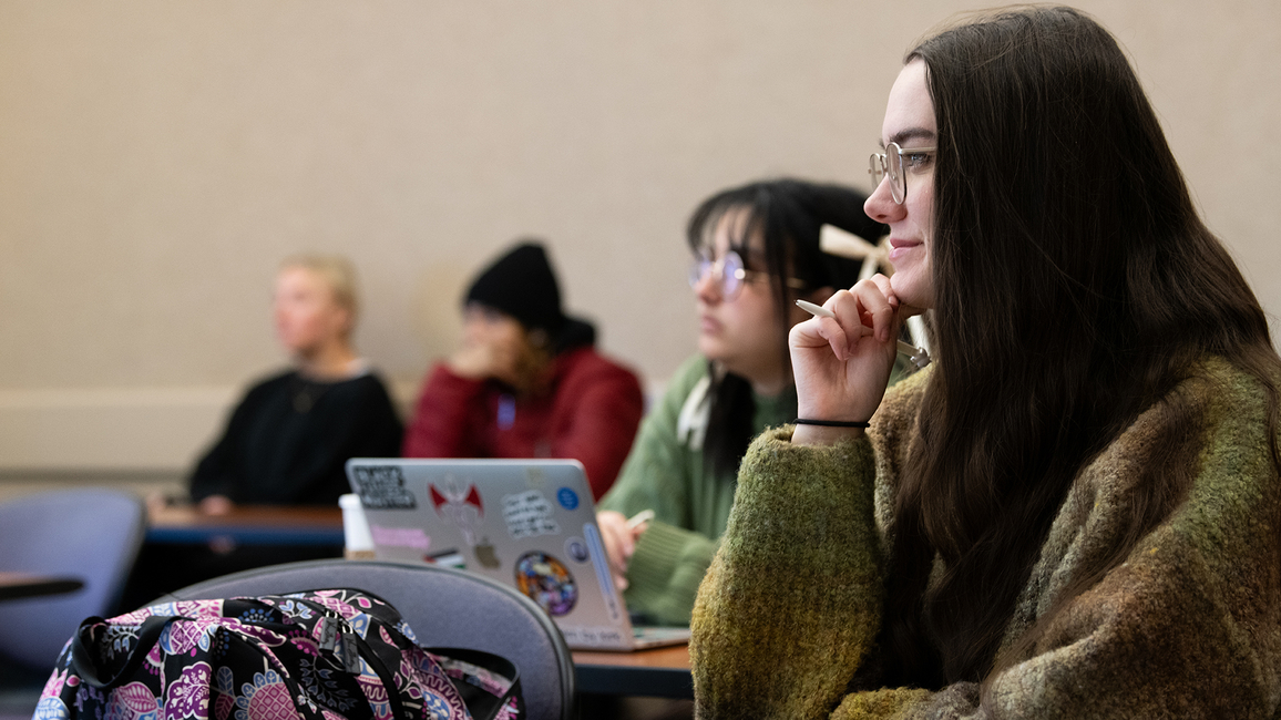 A student in class smiling with a notebook on the desk and pen in hand.