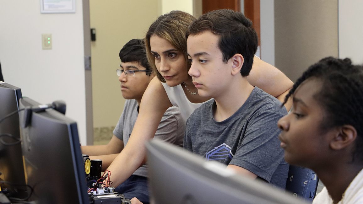 Elham Buxton (center) is shown reaching past a student to control a computer during an AI summer camp.