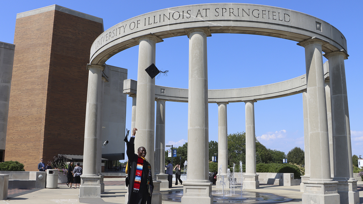 A student in cap and gown standing in front of the colonnade throws his hat into the air. 