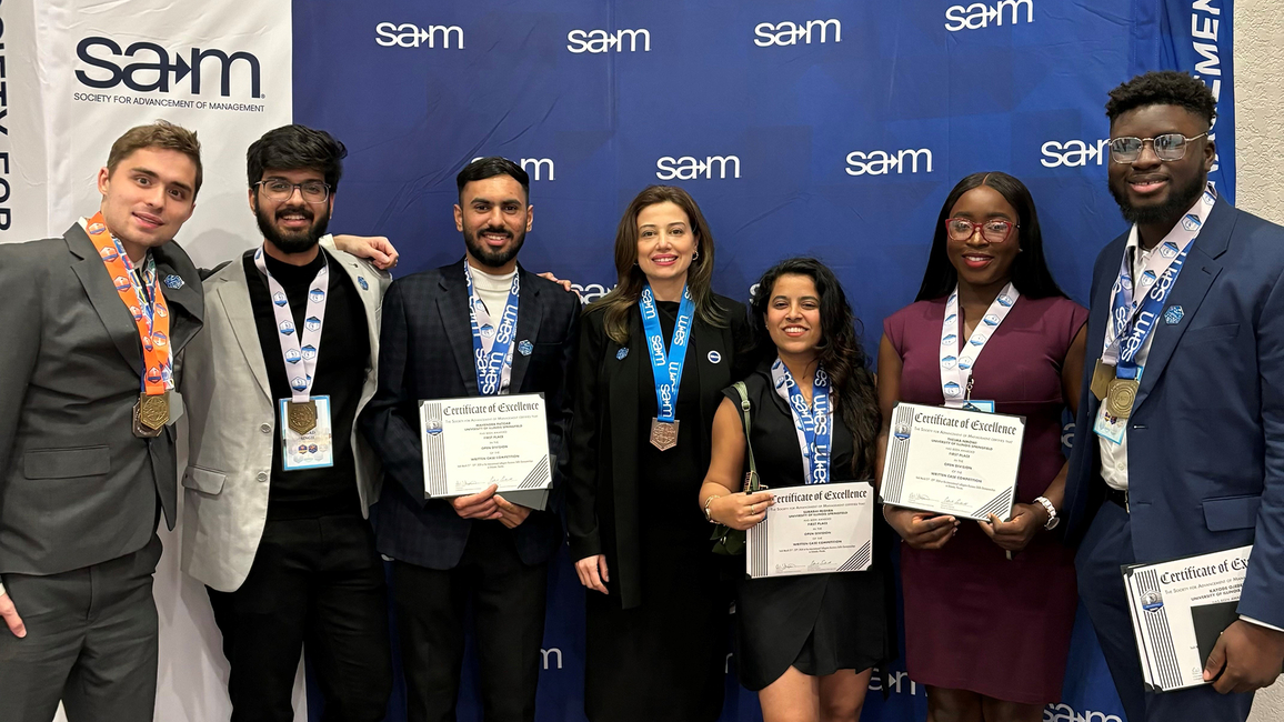 A group of six students and a faculty member posing for a photo, while holding awards, in front of a SAM banner.