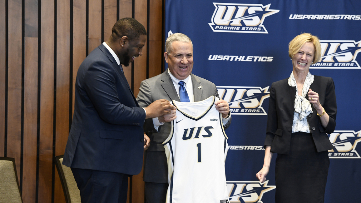 Mike Hermann holding a basketball jersey reading "UIS 1" stands next to Vice Chancellor Jamarco Clark and Chancellor Janet Gooch laughing on stage.