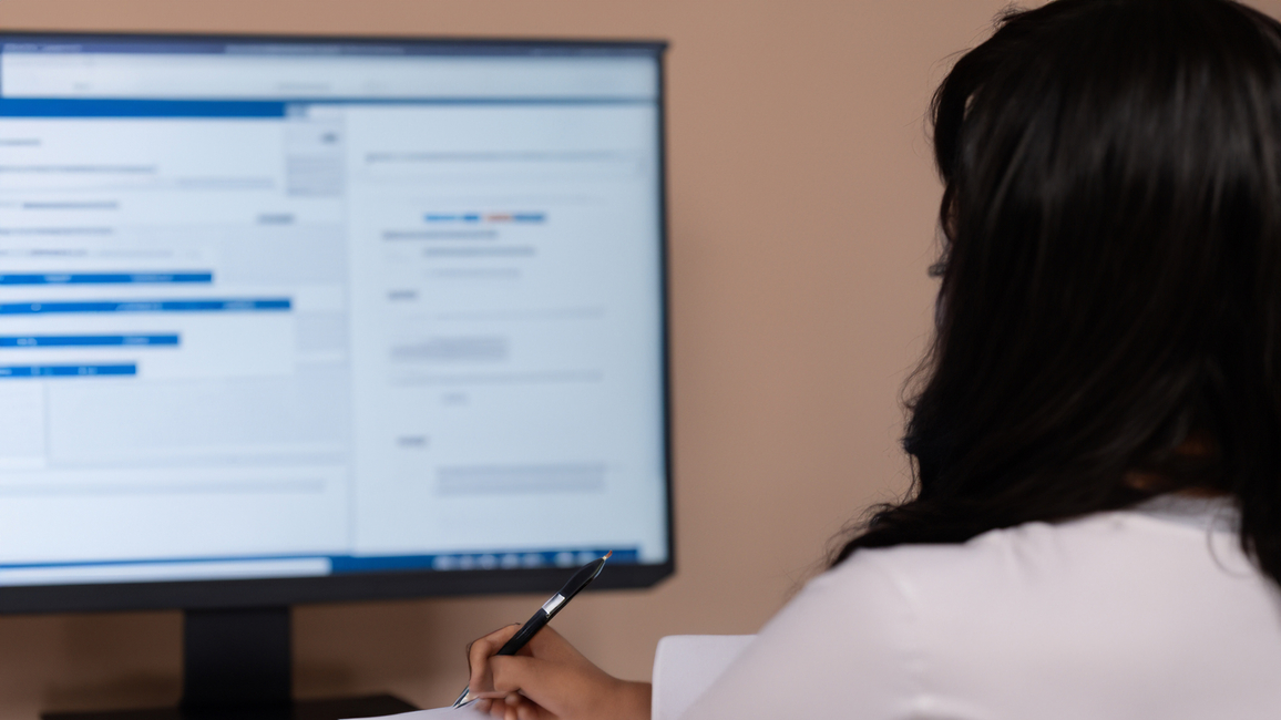 A person from behind sitting at a desk and holding a pen in front of a blurred computer screen.