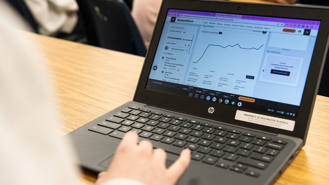 A laptop with stock trading information on the screen sitting on a desk with a person's hand above the mousepad. 