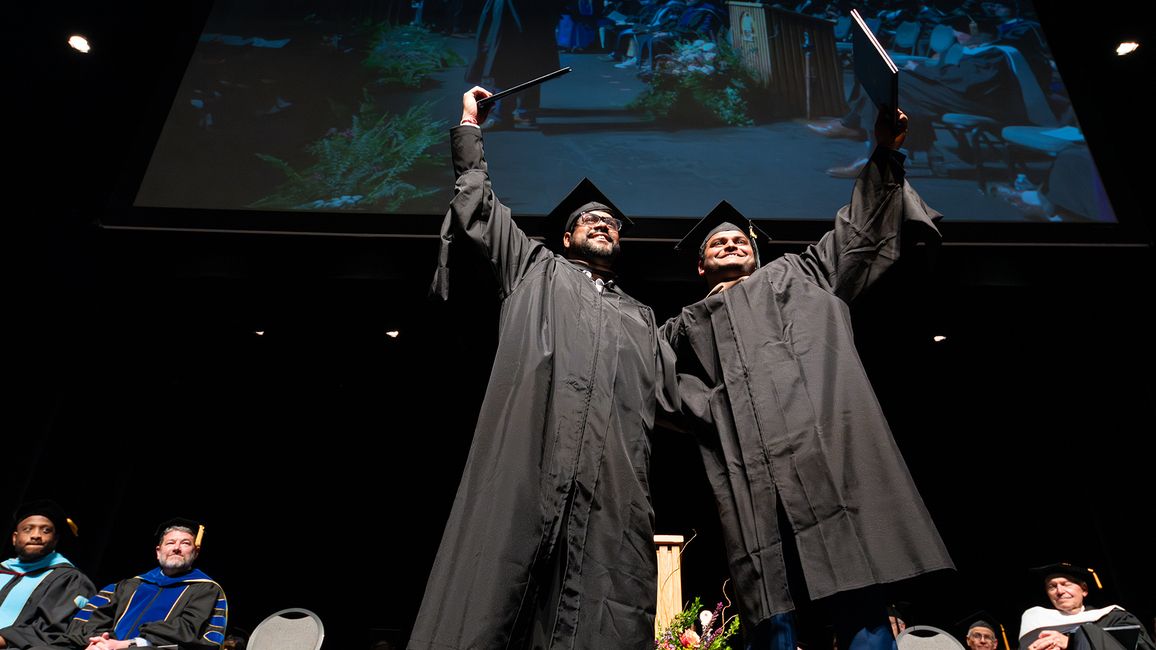 Two graduates in black caps and gowns celebrate on stage by raising their diplomas triumphantly, smiling as they stand side by side. Behind them, faculty members in academic regalia sit in rows and a large screen displays a projection of the moment above them.