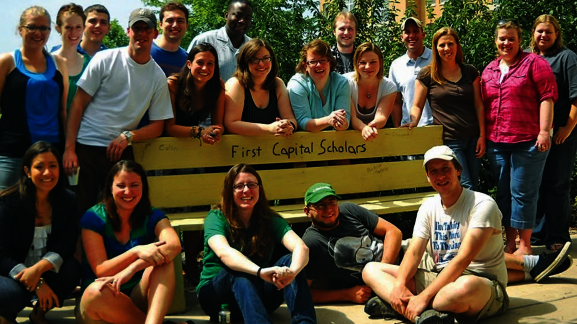 Students gather around a bench that says, "First Capital Scholars."