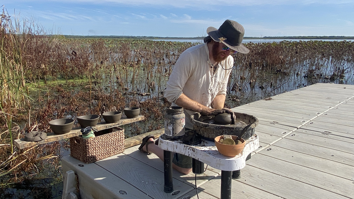 A person shapes clay on a pottery wheel outdoors on a dock at the Emiquon Preserve, surrounded by wetland plants and water.