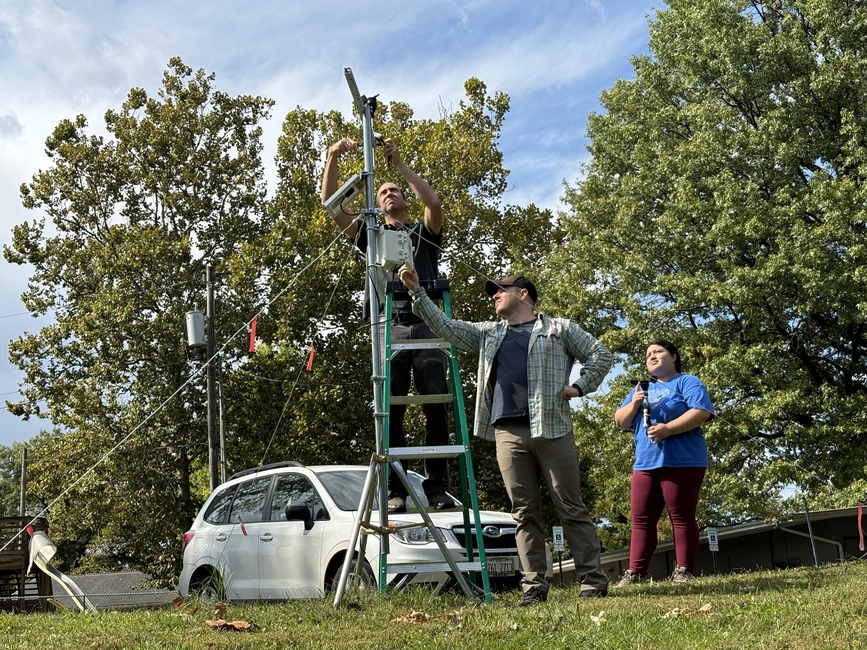 Three researchers assembling weather monitoring station on ladder with atmospheric sensors in parking area