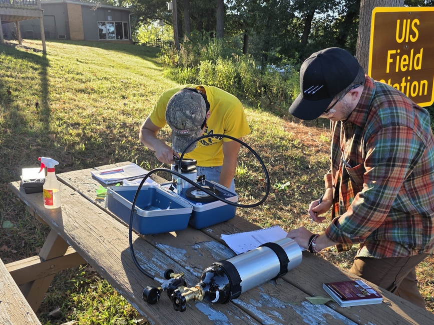 Field scientists operating water quality sensors and documenting plant samples on picnic table at research site