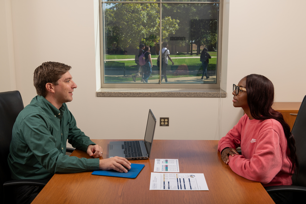 Two people sitting across a desk in an office meeting, laptop and papers on the table.