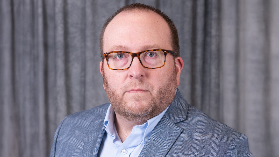 Man with glasses in a blue plaid suit, serious expression, gray background.