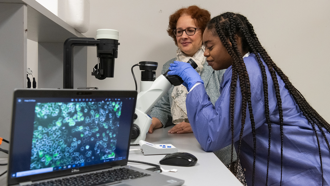 A student in a lab coat looks through a microscope while a professor observes beside her, with cell images displayed on a nearby laptop.