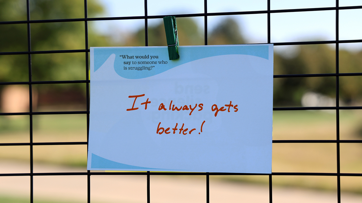 Note with "It always gets better!" clipped to a fence, blurry campus in background.