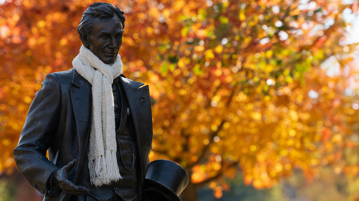 the lincoln statue on the quad has a real scarf wrapped around his neck and there are autumnal leaves on a tree in the background
